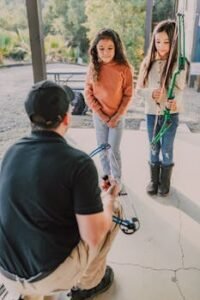 Two young girls learning archery from an instructor with a compound bow outdoors.
