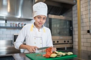 A young chef in a professional kitchen slicing vegetables on a cutting board.